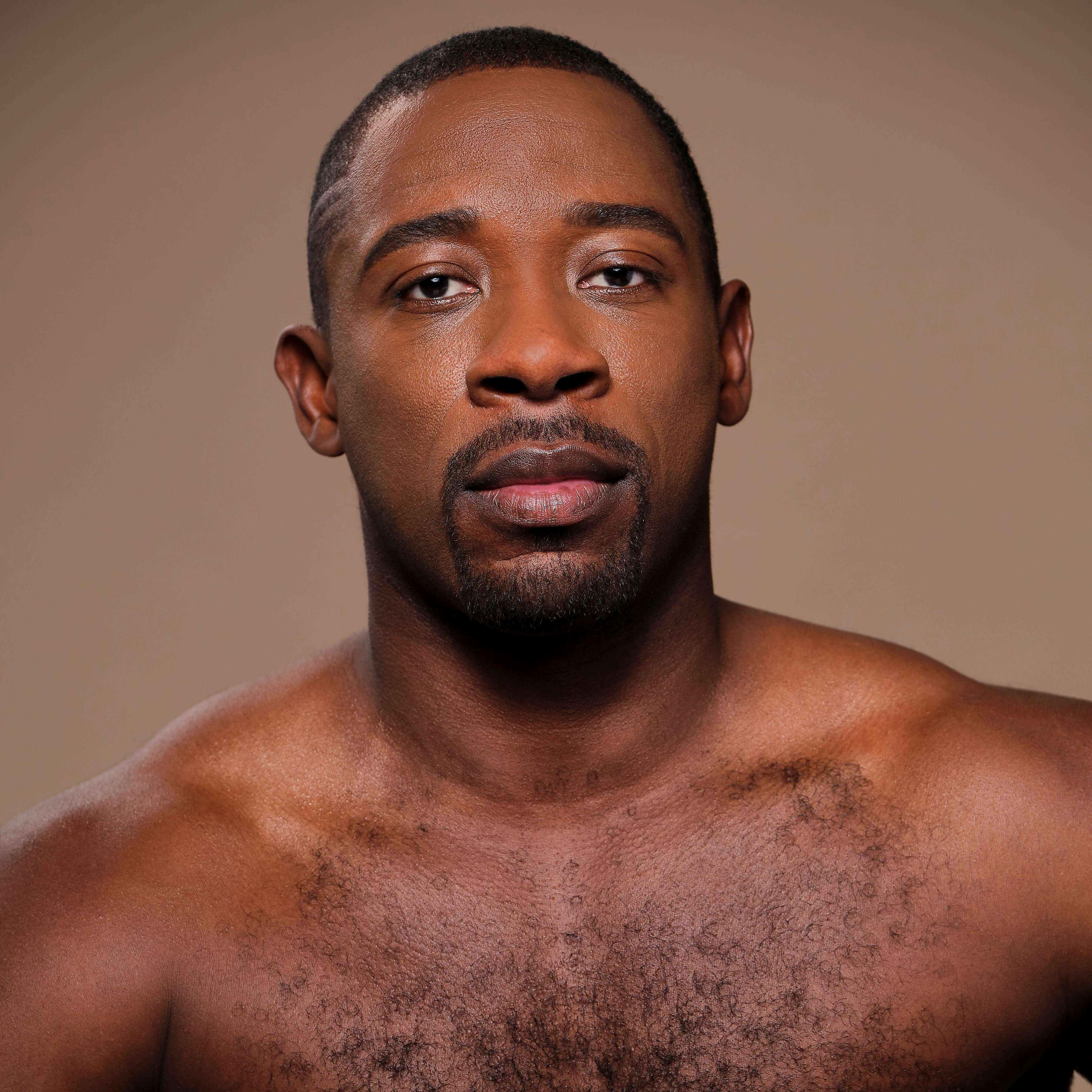 Image Description: Iquail, a Black man with a buzz cut and goatee, looks directly into the camera. He is bare-chested, set against a light brown background